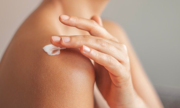 Nahaufnahme einer Schulter; eine Hand trägt mit den Fingerspitzen eine kleine Menge Creme auf die Haut auf.